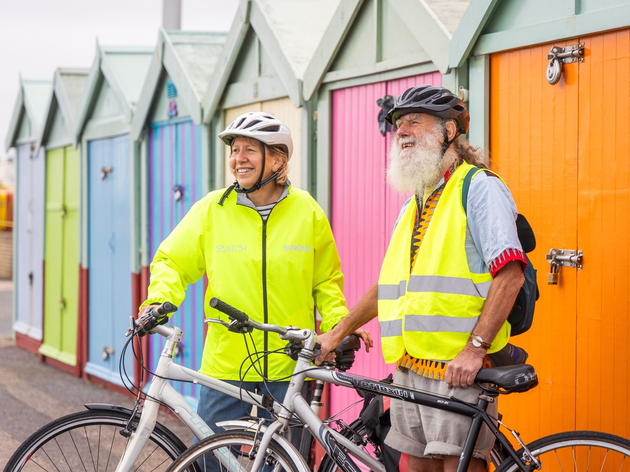 Two older adults standing with bicycles beside colourful beach huts.