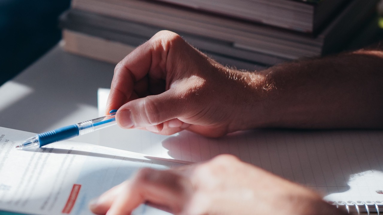 A hand holds a pen above papers beside an open notebook.