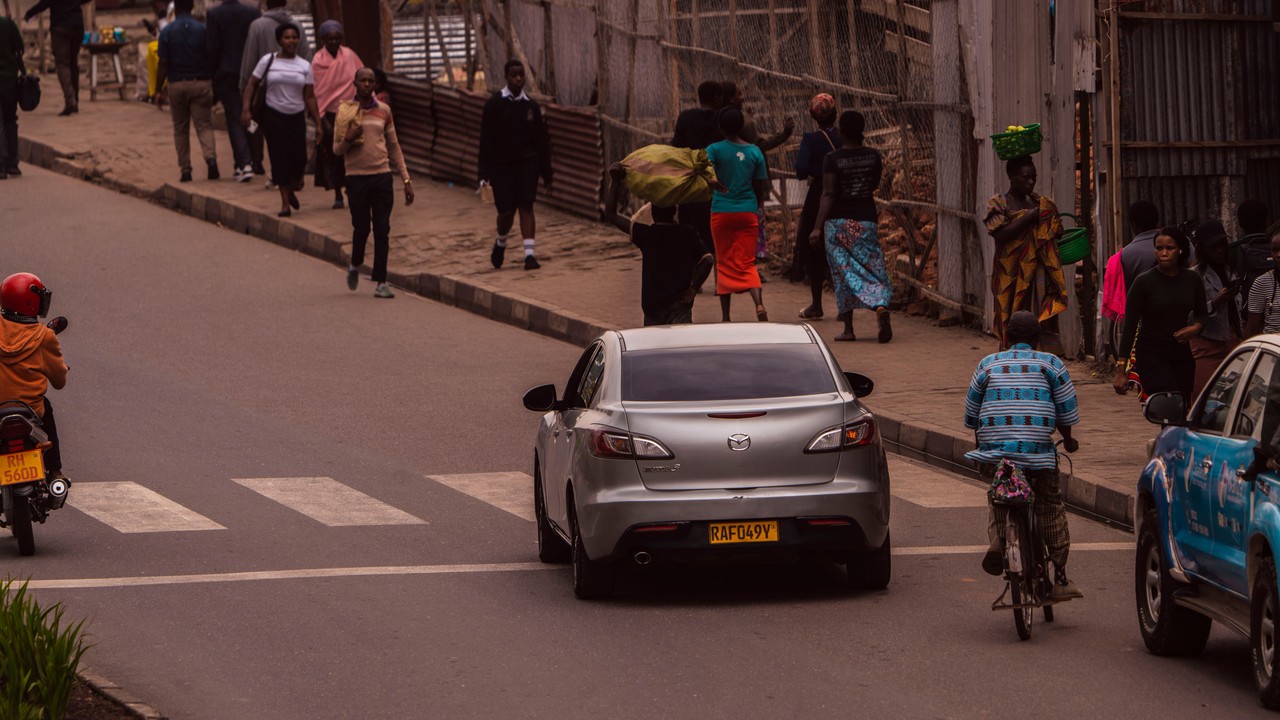 Cars and motorbikes travel along a street as people walk beside the road.